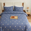 A blue bedding set with a repeating white heart pattern, styled on a bed with a wooden headboard, white pillows, and a small tray of mugs on top.