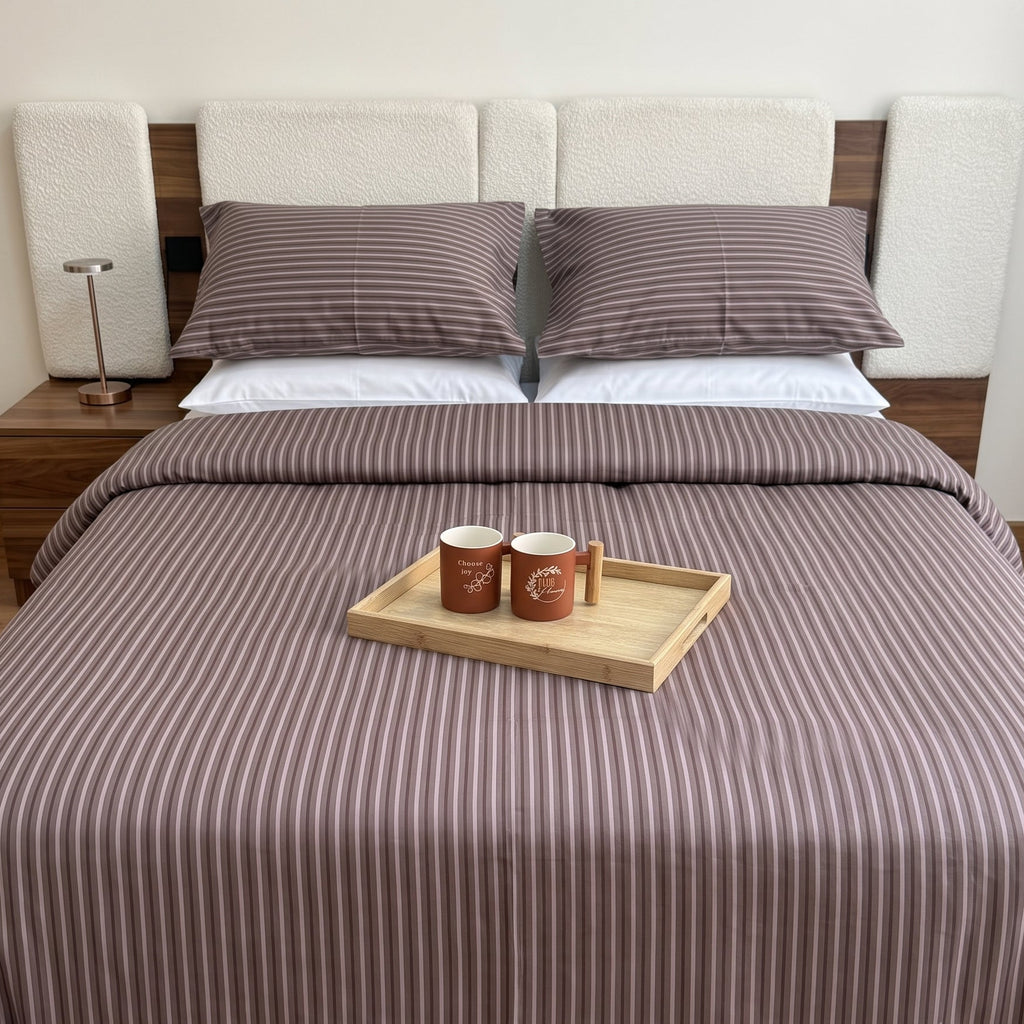 A neatly made bed in a bedroom setting, featuring a brown and white vertically striped bedding set with matching pillows and a duvet cover. A light wood tray with two mugs sits on top of the bed.