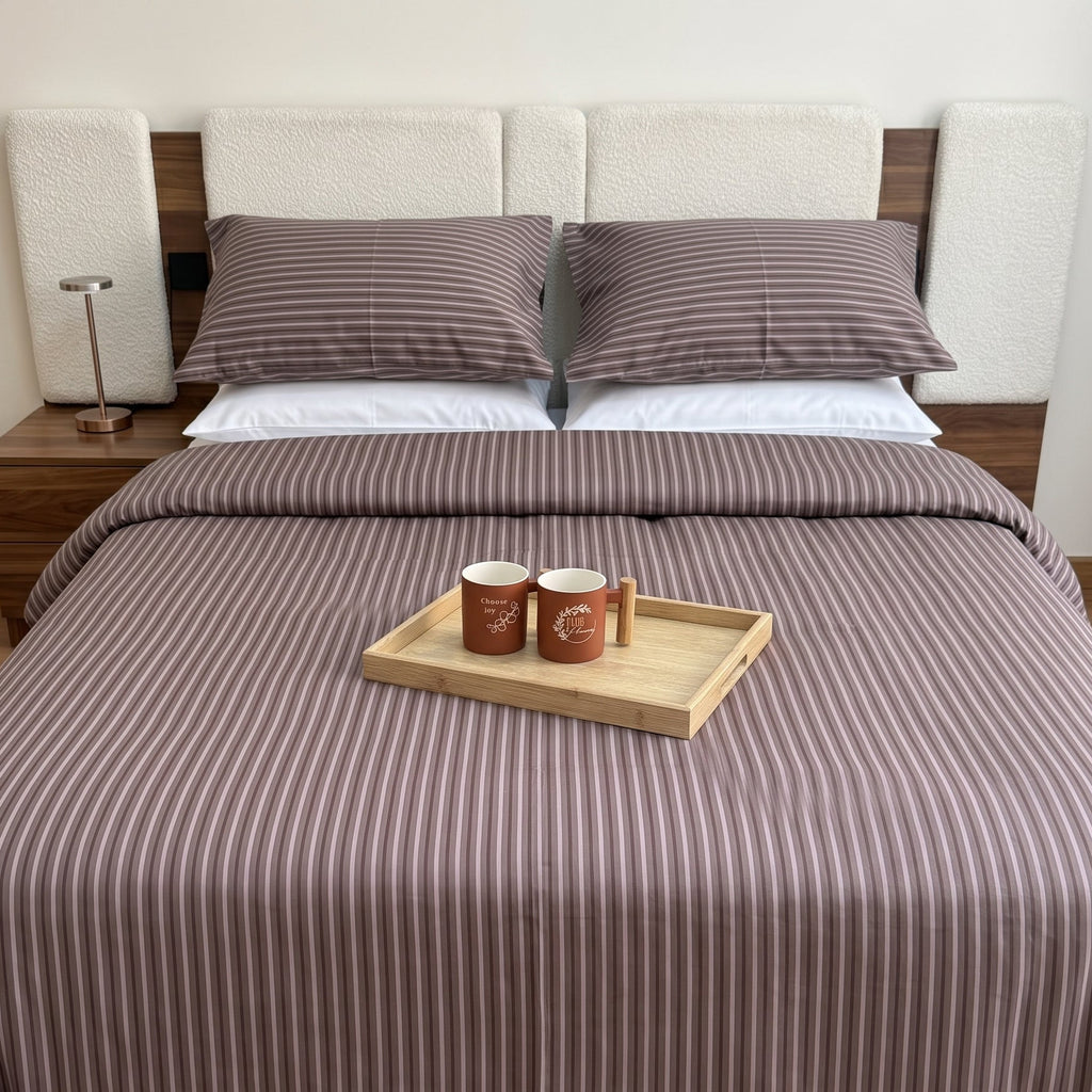 A neatly made bed in a bedroom setting, featuring a brown and white vertically striped bedding set with matching pillows and a duvet cover. A light wood tray with two mugs sits on top of the bed.