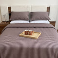 A neatly made bed in a bedroom setting, featuring a brown and white vertically striped bedding set with matching pillows and a duvet cover. A light wood tray with two mugs sits on top of the bed.
