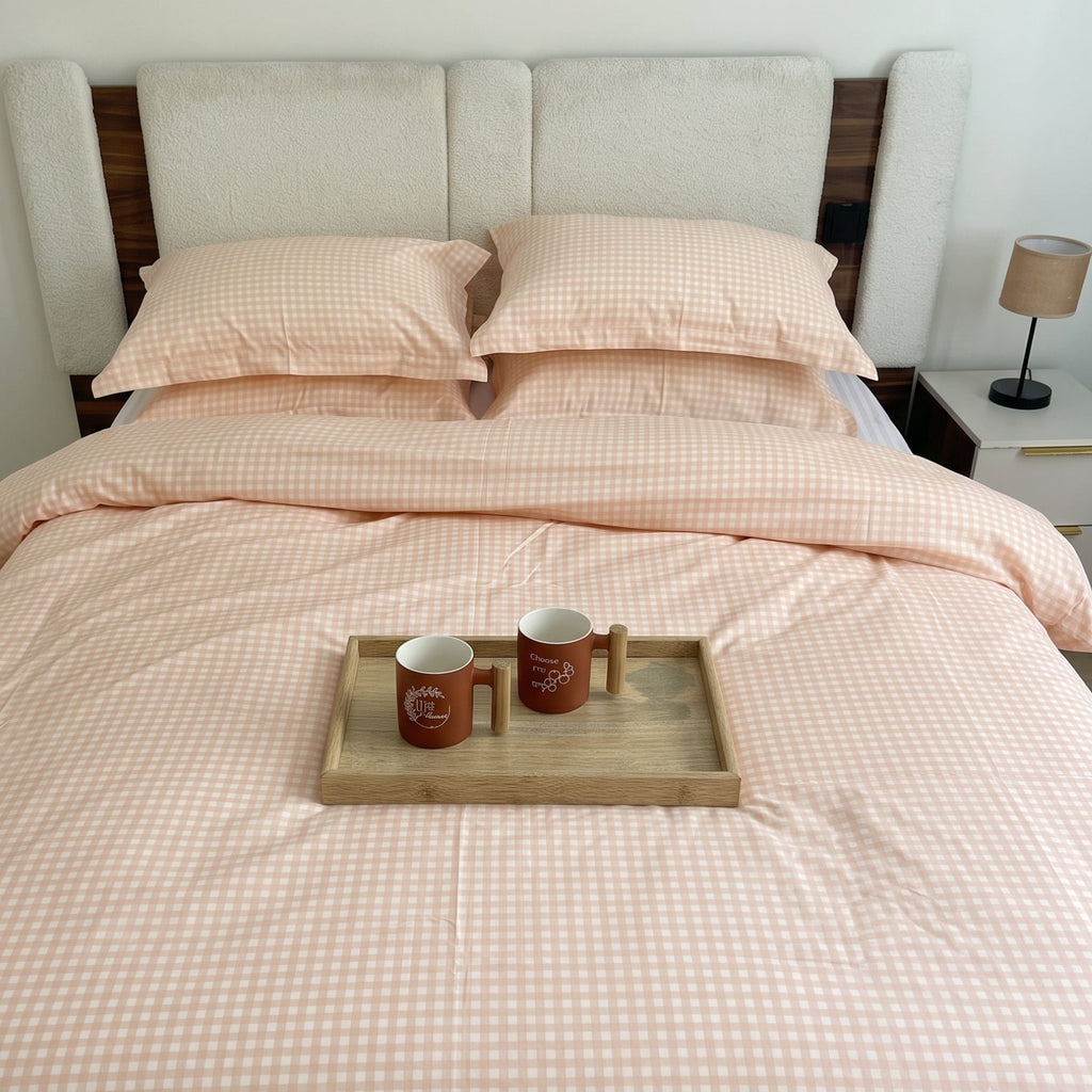 A neatly made bed with a pale blush pink and white gingham check duvet cover and matching pillowcases, styled in a bedroom with a wooden headboard, bedside lamp, and a small tray holding two mugs on the bed.