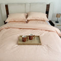 A neatly made bed with a pale blush pink and white gingham check duvet cover and matching pillowcases, styled in a bedroom with a wooden headboard, bedside lamp, and a small tray holding two mugs on the bed.