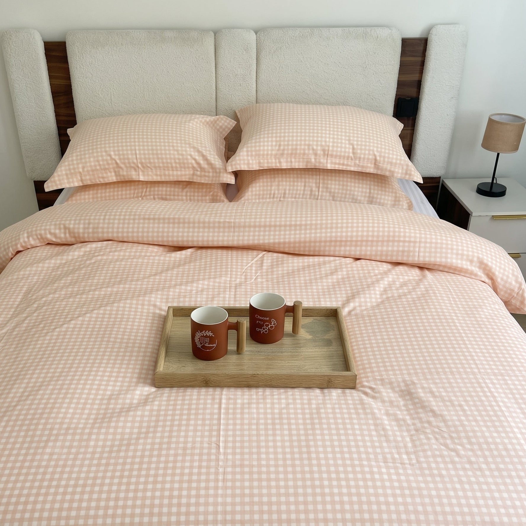 A neatly made bed with a pale blush pink and white gingham check duvet cover and matching pillowcases, styled in a bedroom with a wooden headboard, bedside lamp, and a small tray holding two mugs on the bed.