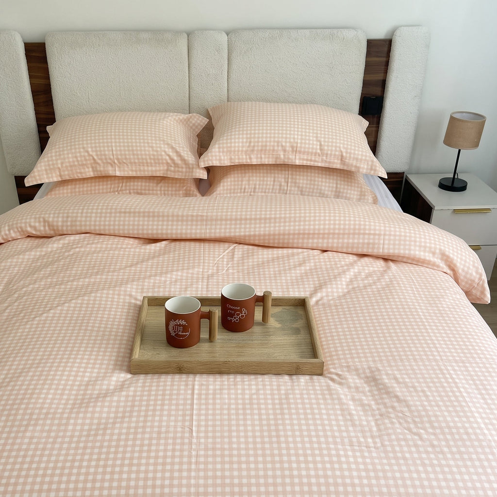 A neatly made bed with a pale blush pink and white gingham check duvet cover and matching pillowcases, styled in a bedroom with a wooden headboard, bedside lamp, and a small tray holding two mugs on the bed.
