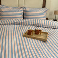 A bed dressed in a striped bedding set with matching pillowcases, featuring vertical stripes in light blue, peach, and white. A small wooden tray with two brown mugs sits on the duvet.