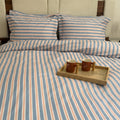 A bed dressed in a striped bedding set with matching pillowcases, featuring vertical stripes in light blue, peach, and white. A small wooden tray with two brown mugs sits on the duvet.