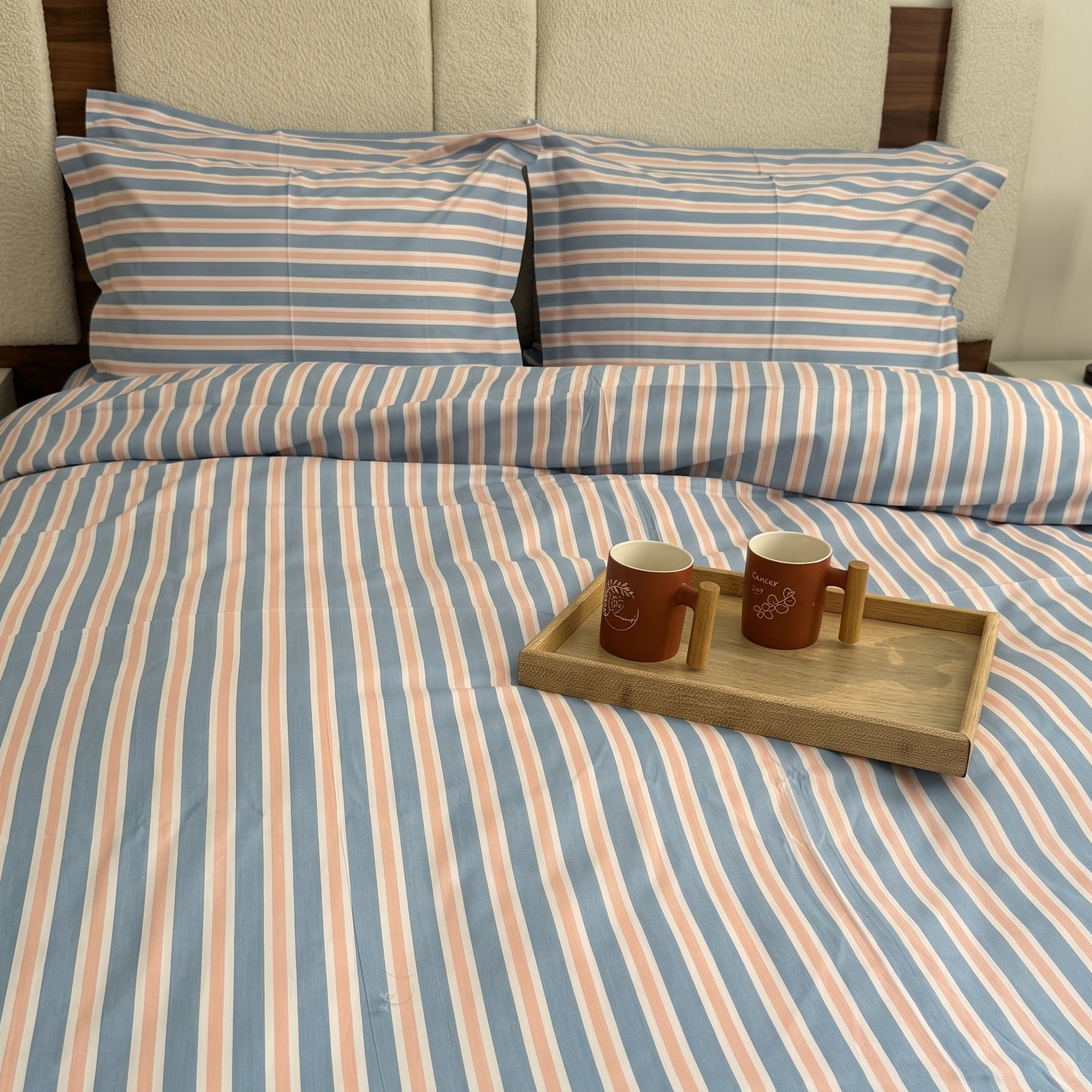 A bed dressed in a striped bedding set with matching pillowcases, featuring vertical stripes in light blue, peach, and white. A small wooden tray with two brown mugs sits on the duvet.