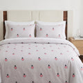A neatly made bed with a white duvet cover and matching pillowcases featuring a repeating pattern of pink strawberries with black seeds and small black dots, set against a beige upholstered headboard and wood floor.