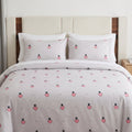 A neatly made bed with a white duvet cover and matching pillowcases featuring a repeating pattern of pink strawberries with black seeds and small black dots, set against a beige upholstered headboard and wood floor.
