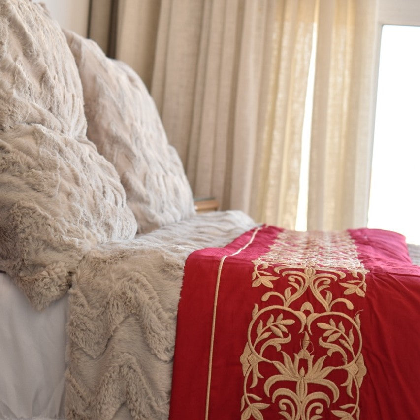 Beige textured blanket draped over a bed with a red and gold patterned throw.