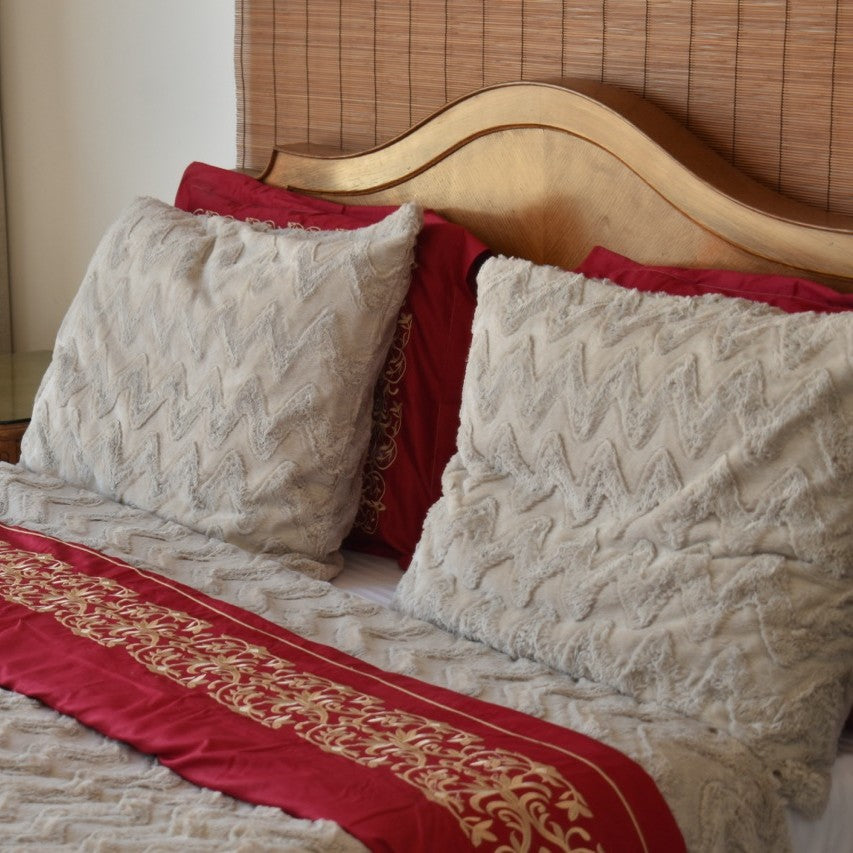Close-up of a bed with textured pillows and a red blanket with gold trim.